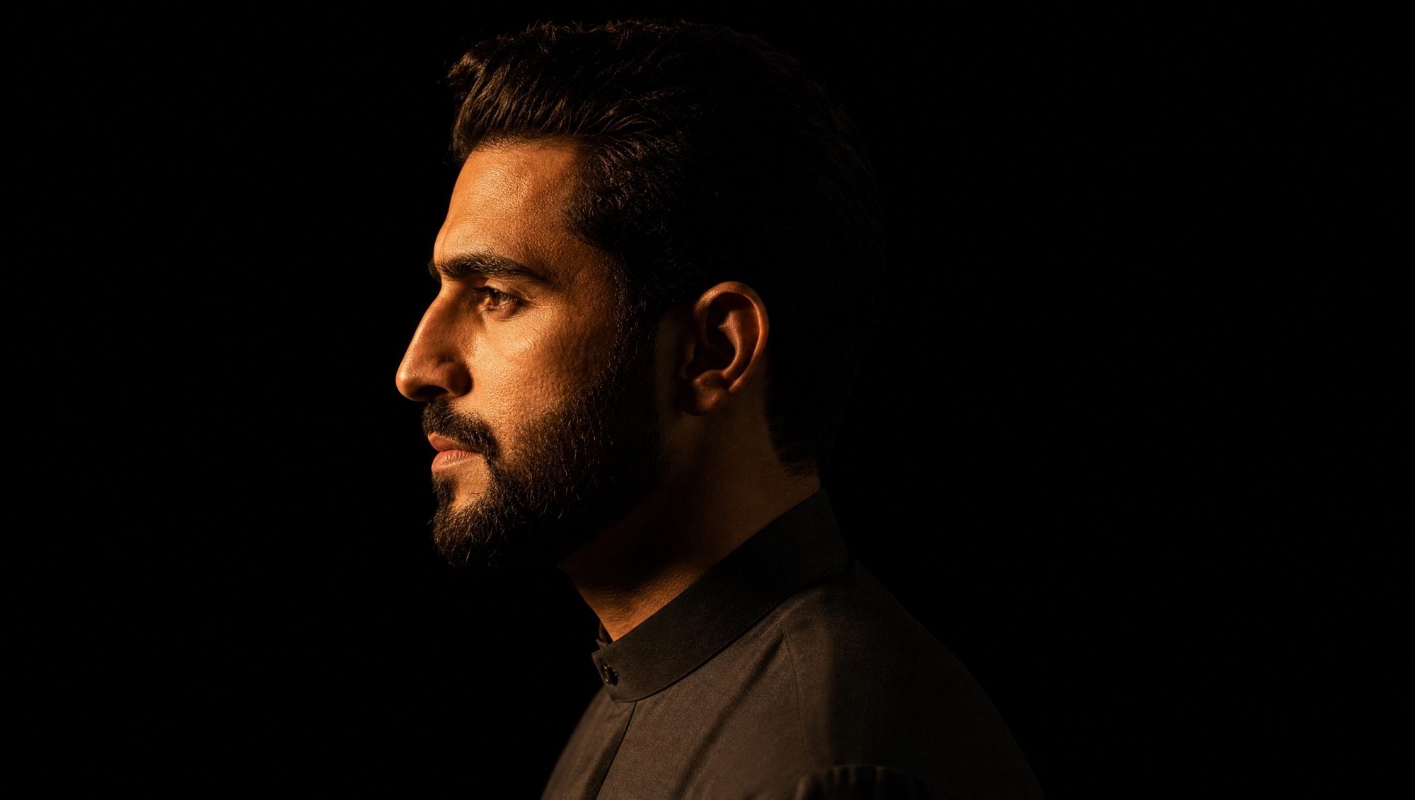 A man in a dark kandura in profile, lit from one side by low sunset light.
