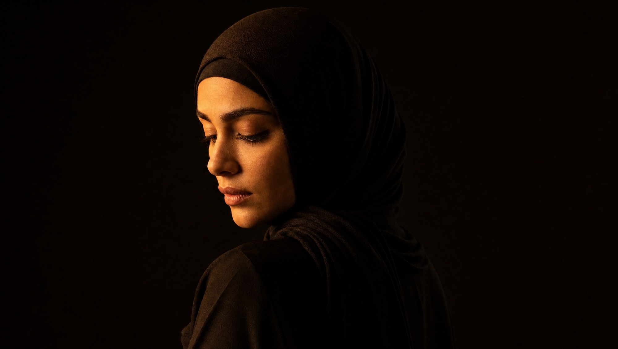 A woman in a black abaya, lit from one side against a dark background.