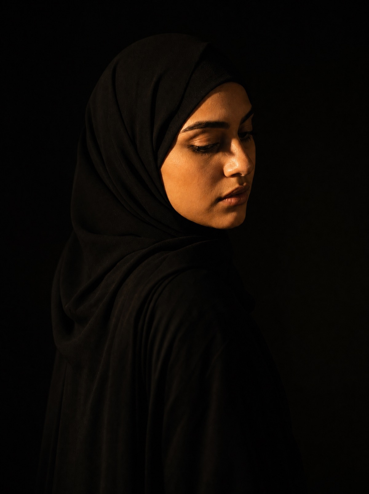 A woman in a black abaya, lit from one side against a dark background.