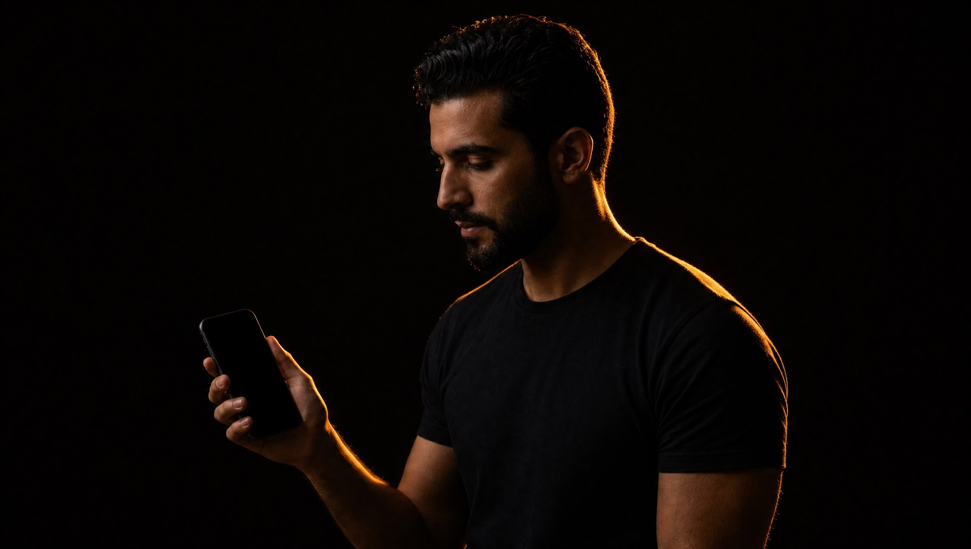 A young man looking at a phone, lit from behind against a dark background.